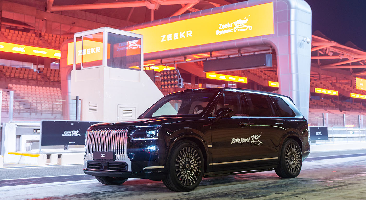 Premium black Zeekr 009 luxury minivan parked on a race track paddock under a large orange Zeekr banner sign.